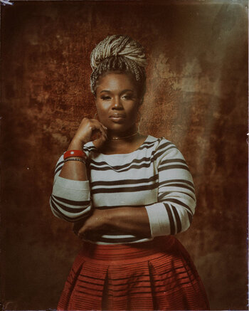 Another portrait against a worn wall. This is of a Black woman with a braided crown of bleached hair and highlights of red. St Dymphna.